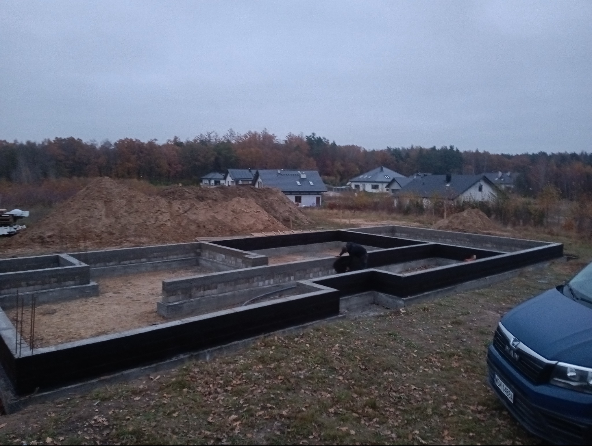 Wykopany i zabezpieczony fundament pod budowę domu jednorodzinnego, widoczny pracownik przy pracy, w tle inne domy i hałda ziemi.