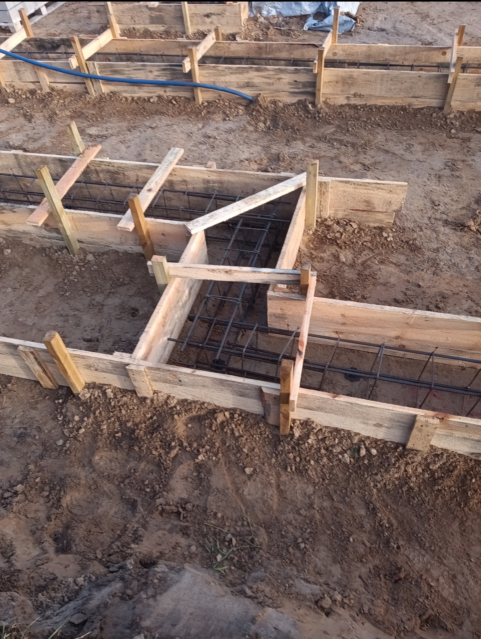 Szczegółowy widok na szalunek fundamentów zbrojonych stalowymi prętami, przygotowany do zalania betonem, w wykopie ziemnym.