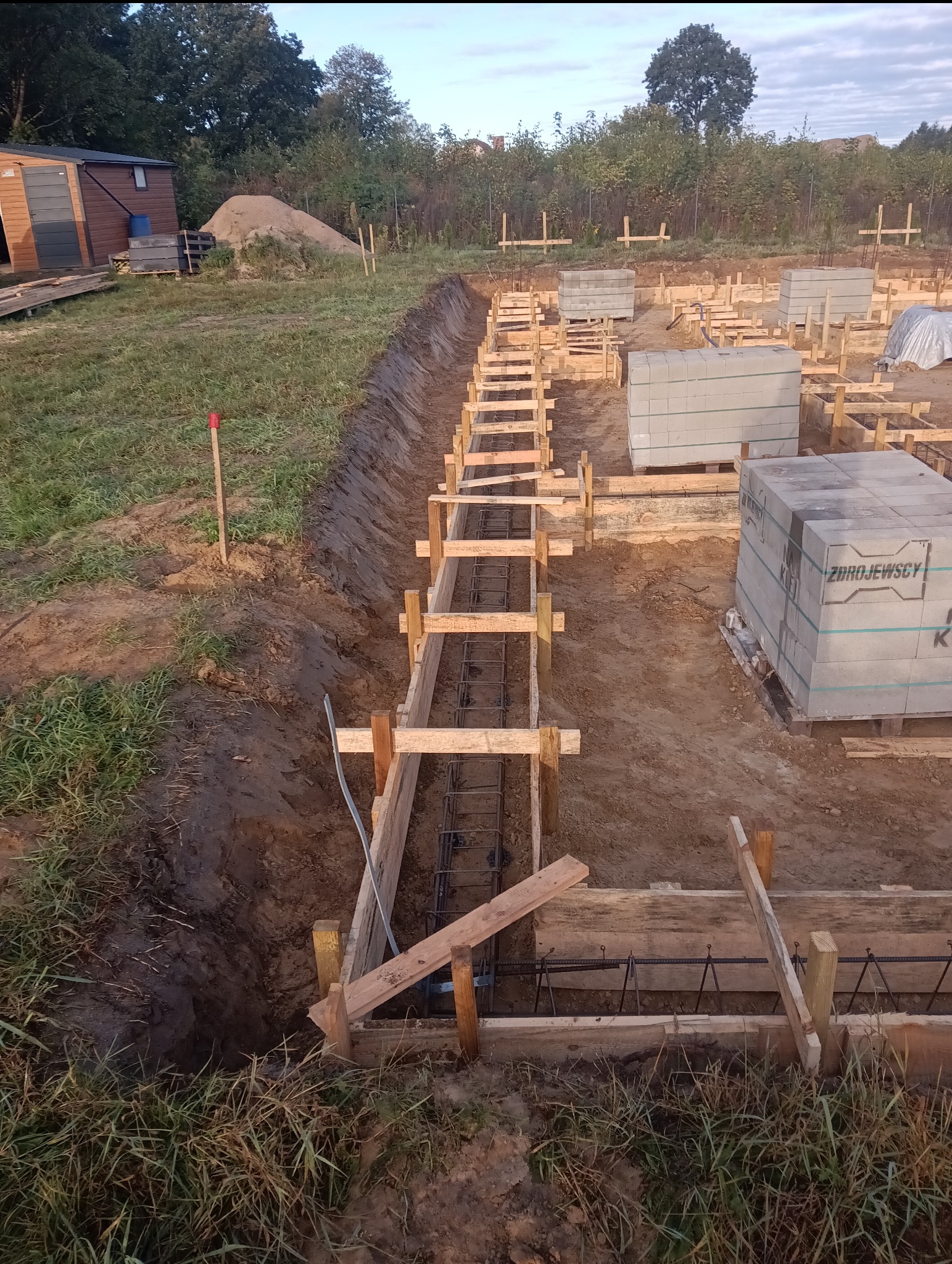 Przygotowanie fundamentów: wykop z drewnianym szalunkiem i zbrojeniem stalowym, obok ułożone bloczki betonowe, w tle sterta piasku i drewniany domek narzędziowy.