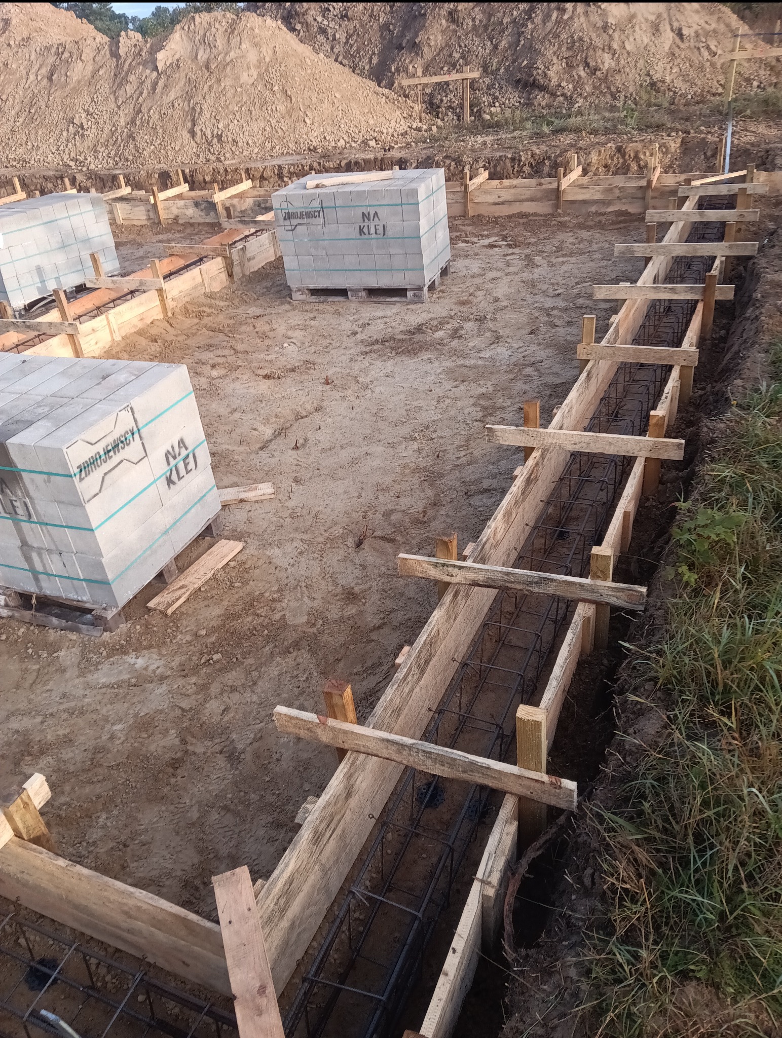 Przygotowanie szalunku fundamentów zbrojonych stalą, widoczne bloczki betonowe na paletach i usypana ziemia w tle.