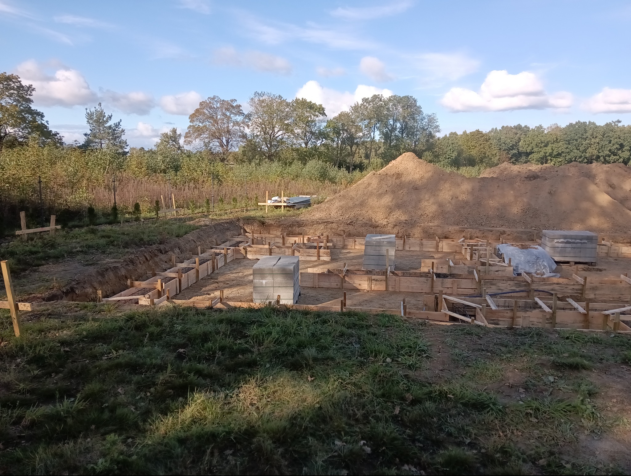 Przygotowanie szalunku fundamentów pod budowę domu, widoczne drewniane konstrukcje, wykop, sterta ziemi oraz bloczki betonowe na placu budowy w słoneczny dzień.