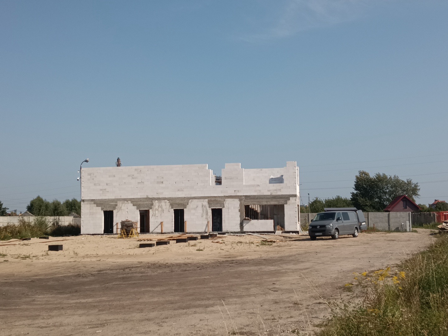 Niedokończony budynek z białych bloczków, widoczne otwory okienne i drzwiowe, palety z materiałami budowlanymi przed budynkiem, szara furgonetka zaparkowana obok, teren budowy.