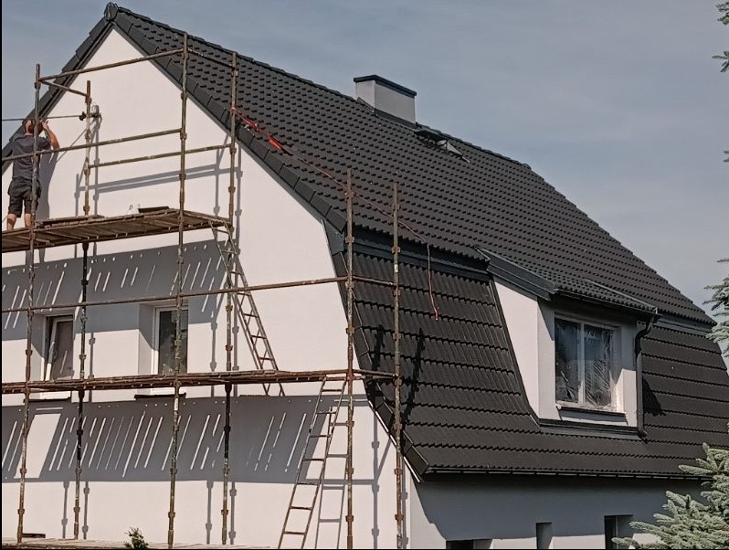 Prace na rusztowaniu przy dwukondygnacyjnym domu z czarną dachówką, malowanie białej elewacji, okno zabezpieczone folią.
