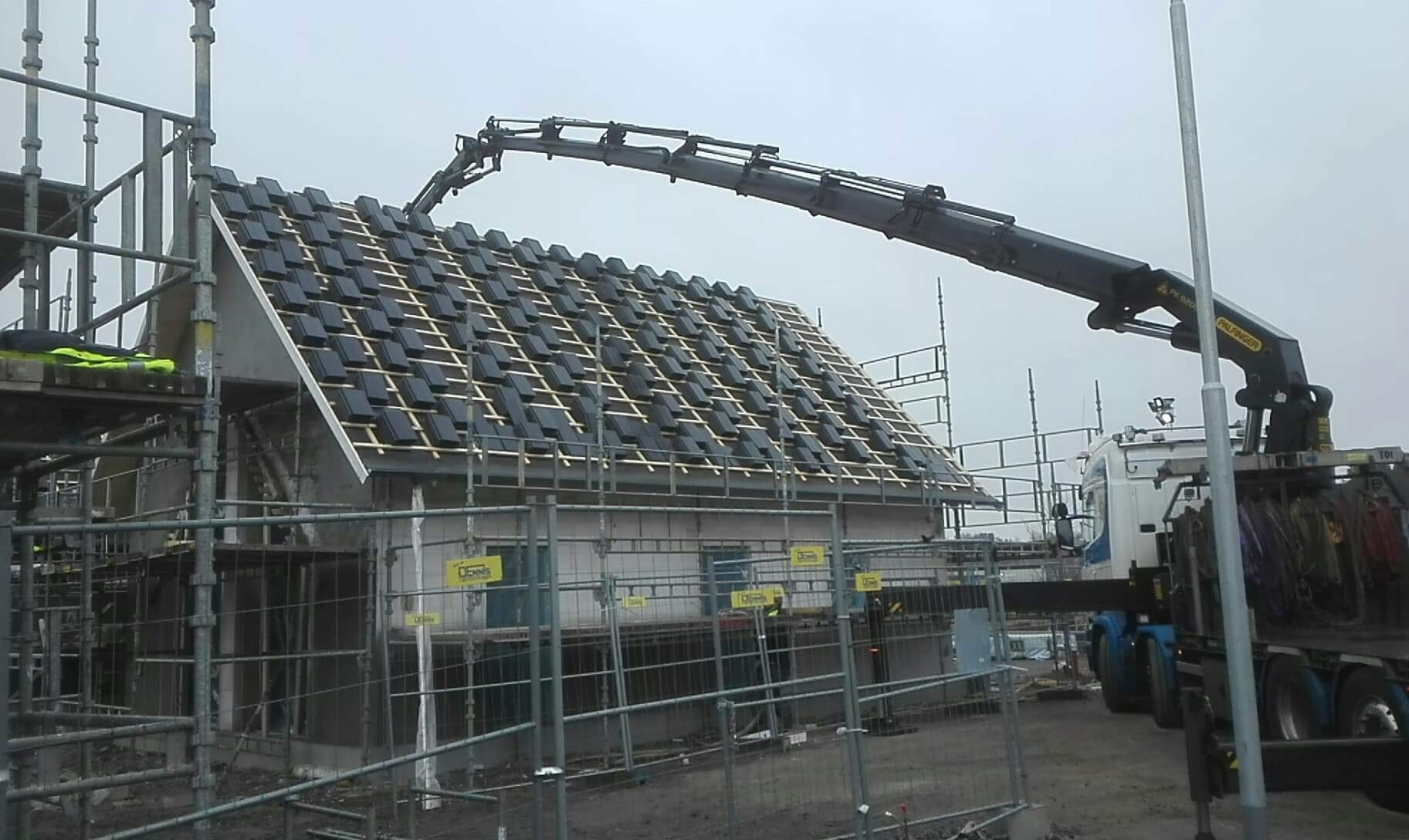 Montaż czarnej dachówki na nowym domu z pomocą dźwigu samochodowego Palfinger, widoczne rusztowania i ogrodzenie placu budowy.