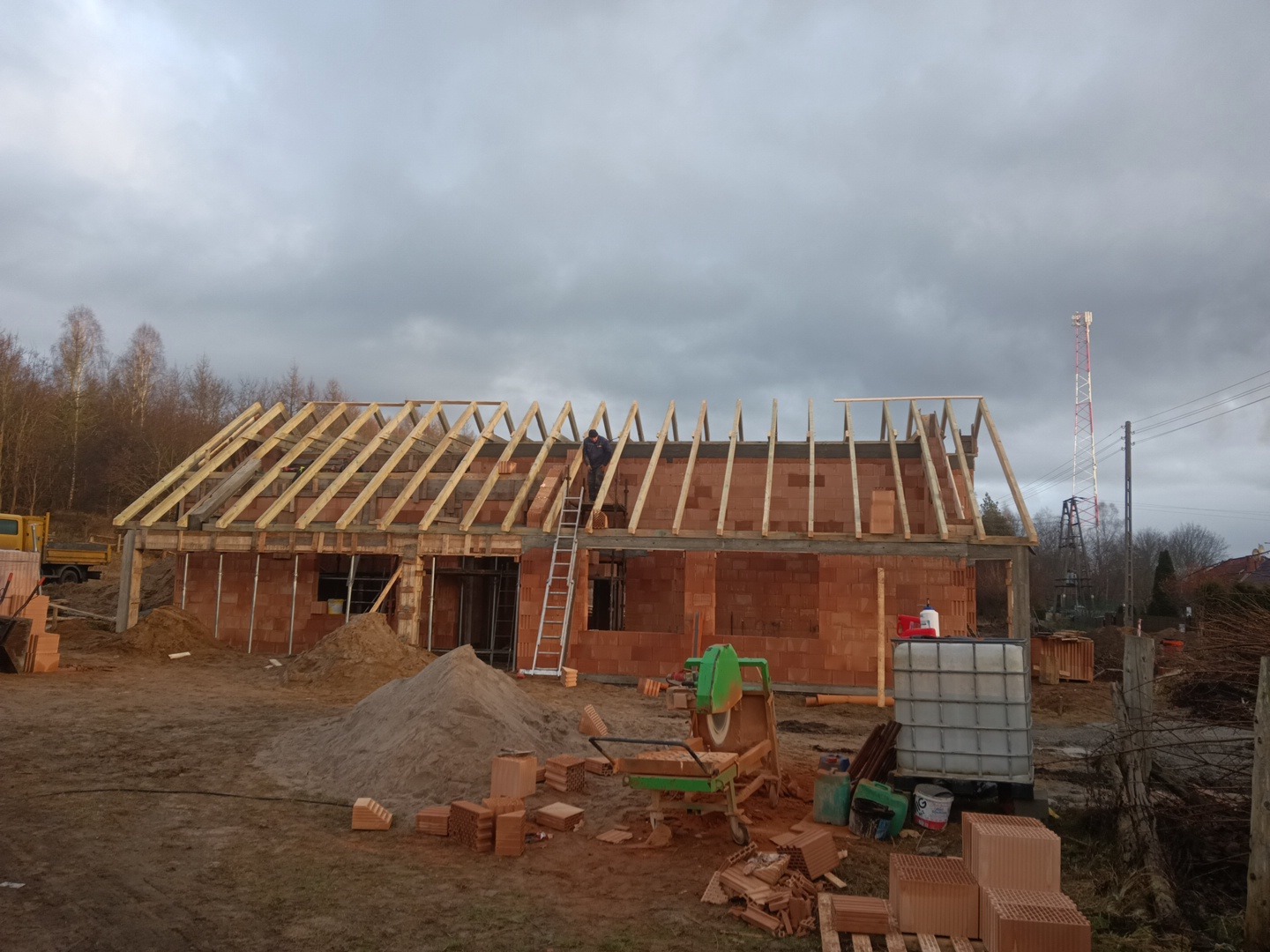 Budowa domu jednorodzinnego: wznoszenie ścian z czerwonej cegły i konstrukcji dachu z drewnianych belek, widoczny robotnik na drabinie, plac budowy z narzędziami i materiałami budowlanymi...