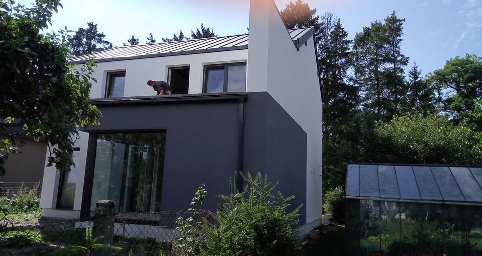 Nowoczesny dom murowany z dwukolorową elewacją, pracownik na dachu przy oknie, widok z poziomu ogrodu z szklarnią w tle.