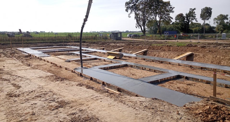Wylewanie betonu do szalunków fundamentowych na placu budowy, widoczna pompa do betonu i przygotowany teren pod budowę.