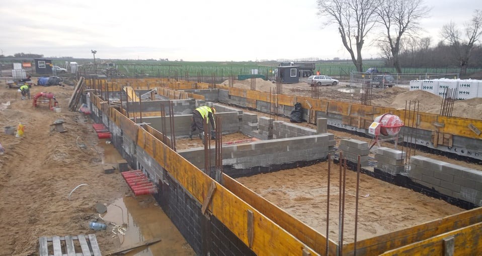 Budowa fundamentów: pracownik w żółtej kamizelce układa bloczki betonowe w szalunkach, widoczne zbrojenia, izolacja, mieszalnik do betonu i teren budowy w tle.