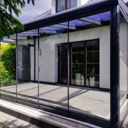 ANGELO Zadaszenia Tarasowe Rolety Żaluzje Ogrody Zimowe Zabudowa Balkonów Moskitiery - Zadaszony taras z metalową konstrukcją i szklanymi ścianami, widoczny z zewnątrz, z fragmentem ogrodu i postacią odbijającą się w szybie.