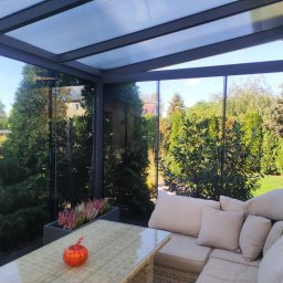 ANGELO Zadaszenia Tarasowe Rolety Żaluzje Ogrody Zimowe Zabudowa Balkonów Moskitiery - Zadaszony taras z przeszklonymi ścianami, widok na zielony ogród, rattanowy stół z dekoracyjną dynią i doniczką z wrzosami, beżowa sofa z poduszkami.