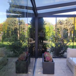 ANGELO Zadaszenia Tarasowe Rolety Żaluzje Ogrody Zimowe Zabudowa Balkonów Moskitiery - Nowoczesna zabudowa tarasowa z przesuwnymi szklanymi panelami, odbijająca w szybach bujną zieleń ogrodu, z widocznym odbiciem osoby robiącej zdjęcie.