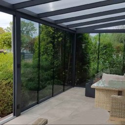 ANGELO Zadaszenia Tarasowe Rolety Żaluzje Ogrody Zimowe Zabudowa Balkonów Moskitiery - Zadaszony taras z przesuwnymi szklanymi panelami, widok na zadbany ogród z trawnikiem i drzewami, rattanowe meble wypoczynkowe z poduszkami.