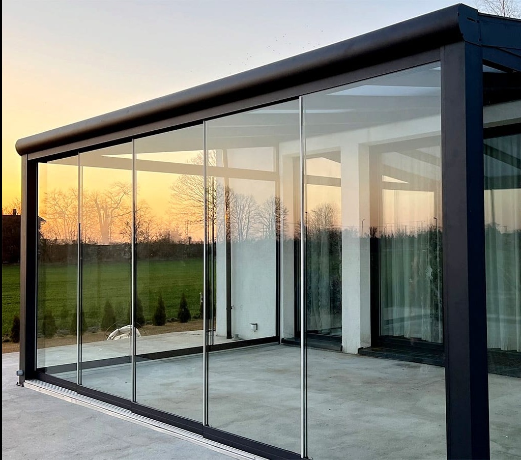Nowoczesna konstrukcja z czarną metalową ramą i przesuwanymi szklanymi panelami, odbijającymi zachodzące słońce i zielony krajobraz, tworząca przeszkloną przestrzeń na tarasie.