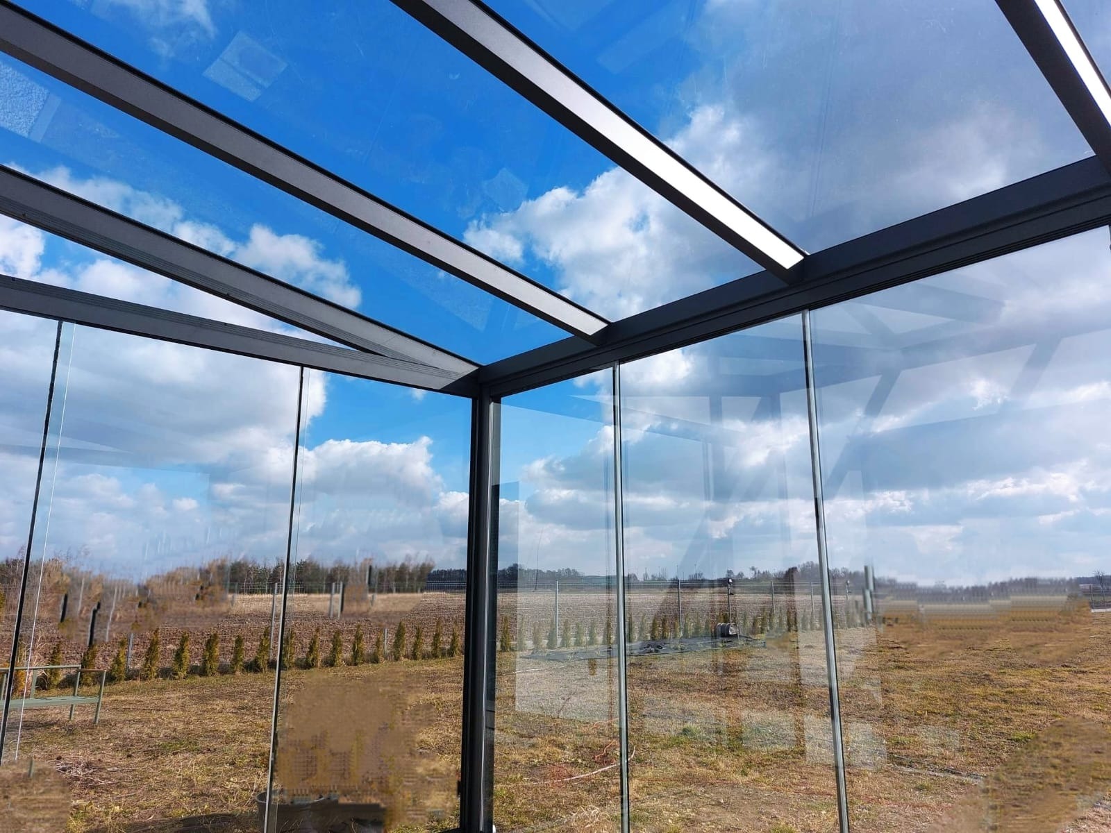 Szklana konstrukcja z metalowym szkieletem, prawdopodobnie oranżeria, z widokiem na pole i błękitne niebo z chmurami odbijającymi się w szybach.