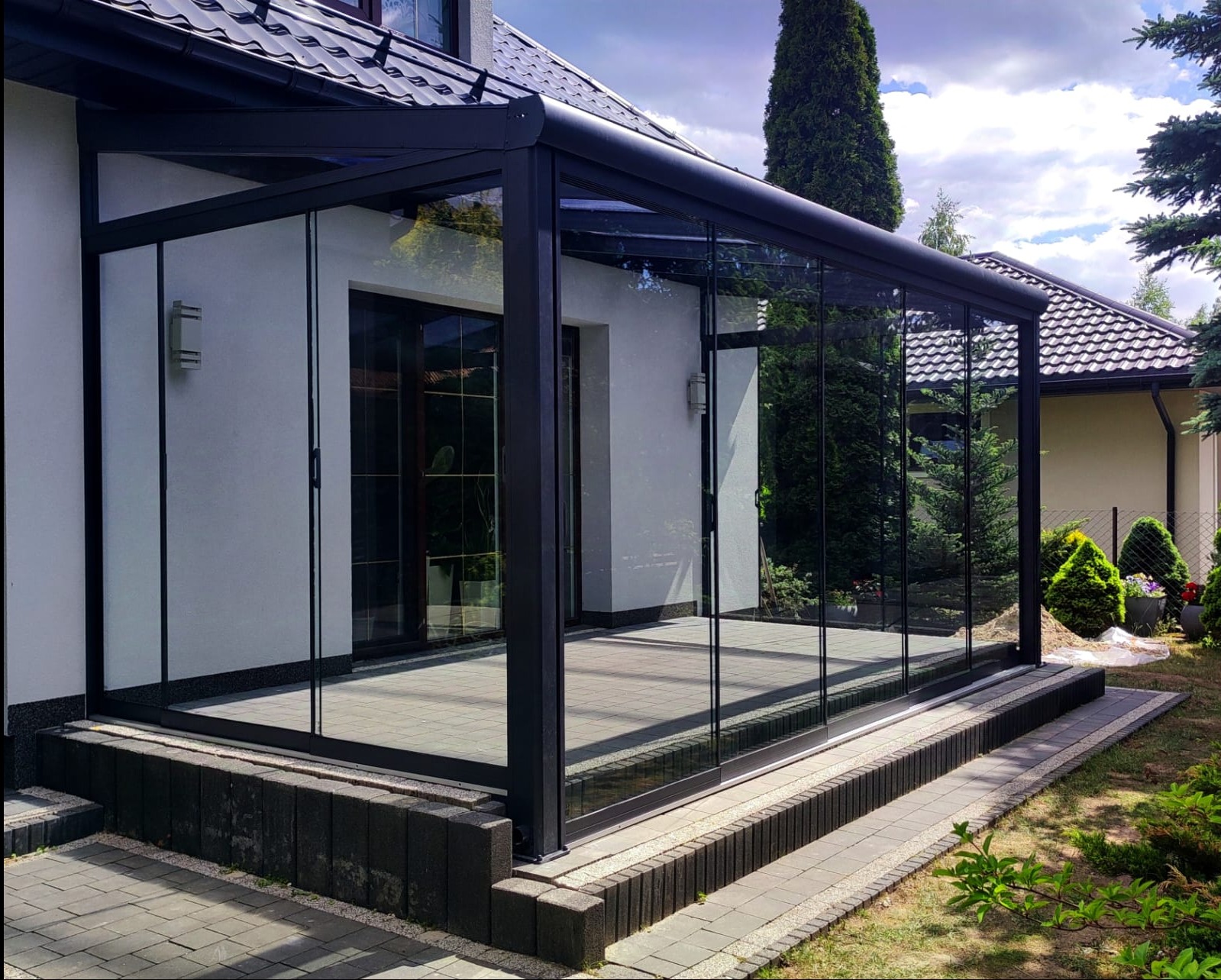 Zadaszony taras z metalową pergolą i szklanymi przesuwnymi ścianami, z widokiem na ogród i kostkę brukową.