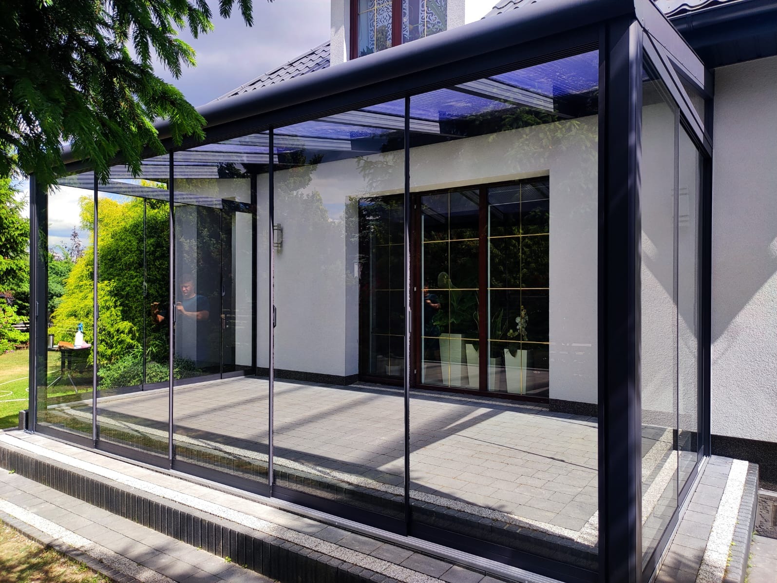 Zadaszony taras z metalową konstrukcją i szklanymi ścianami, widoczny z zewnątrz, z fragmentem ogrodu i postacią odbijającą się w szybie.