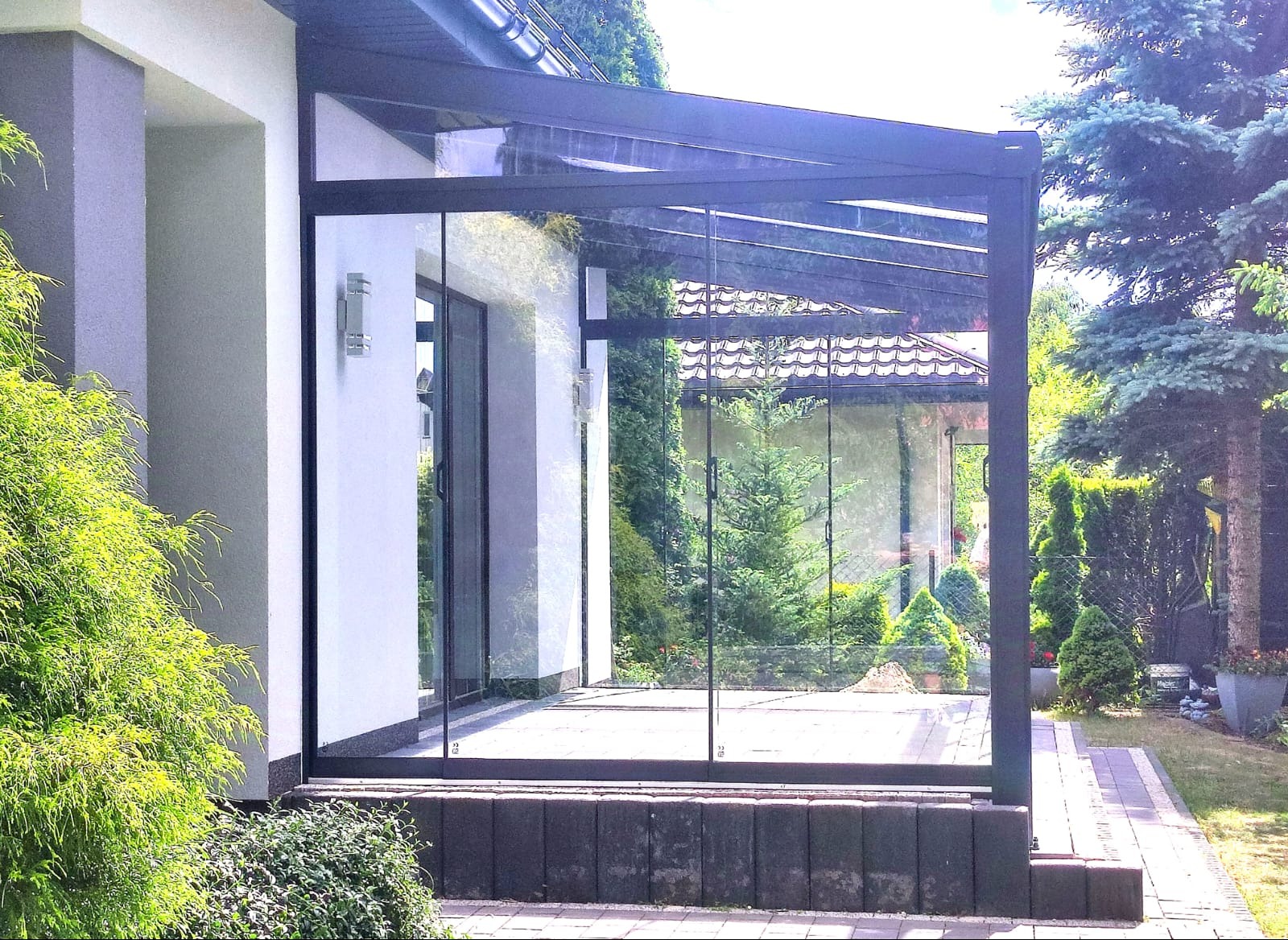 Nowoczesna pergola z czarną metalową konstrukcją i szklanymi ścianami, przylegająca do białego budynku z oświetleniem zewnętrznym, widok na zadbany ogród z iglakami.