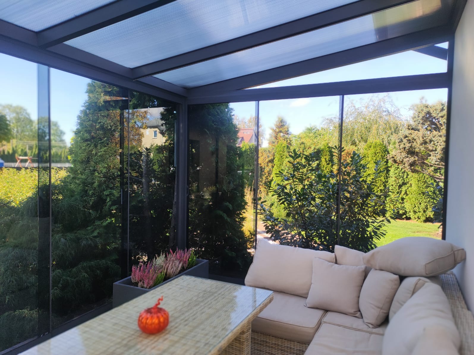 Zadaszony taras z przeszklonymi ścianami, widok na zielony ogród, rattanowy stół z dekoracyjną dynią i doniczką z wrzosami, beżowa sofa z poduszkami.