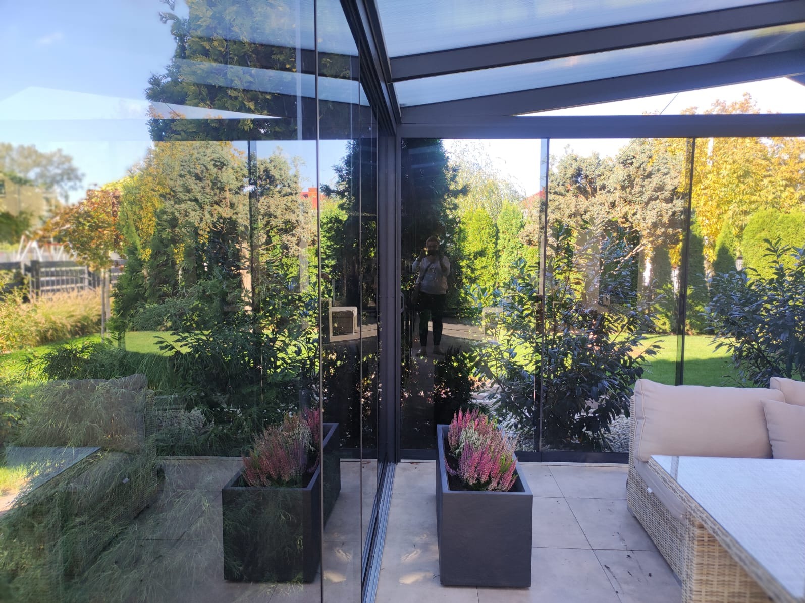Nowoczesna zabudowa tarasowa z przesuwnymi szklanymi panelami, odbijająca w szybach bujną zieleń ogrodu, z widocznym odbiciem osoby robiącej zdjęcie.