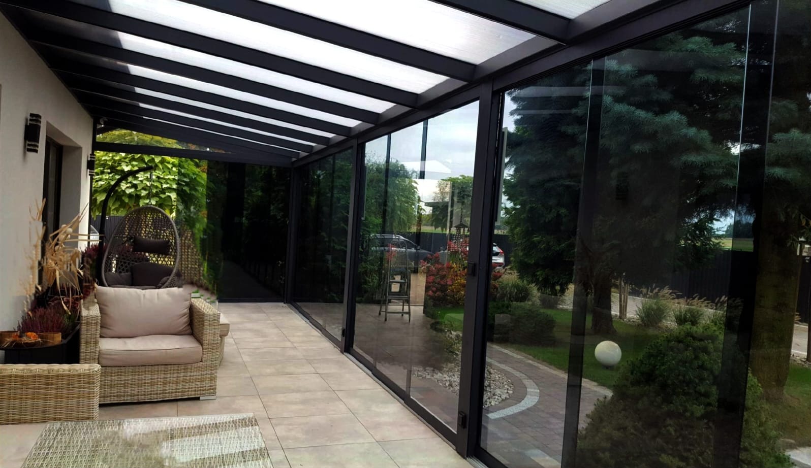 Zadaszony taras z przesuwnymi szklanymi ścianami w Siedlcach, z widokiem na zadbany ogród. Na tarasie rattanowy fotel wiszący i komplet wypoczynkowy.