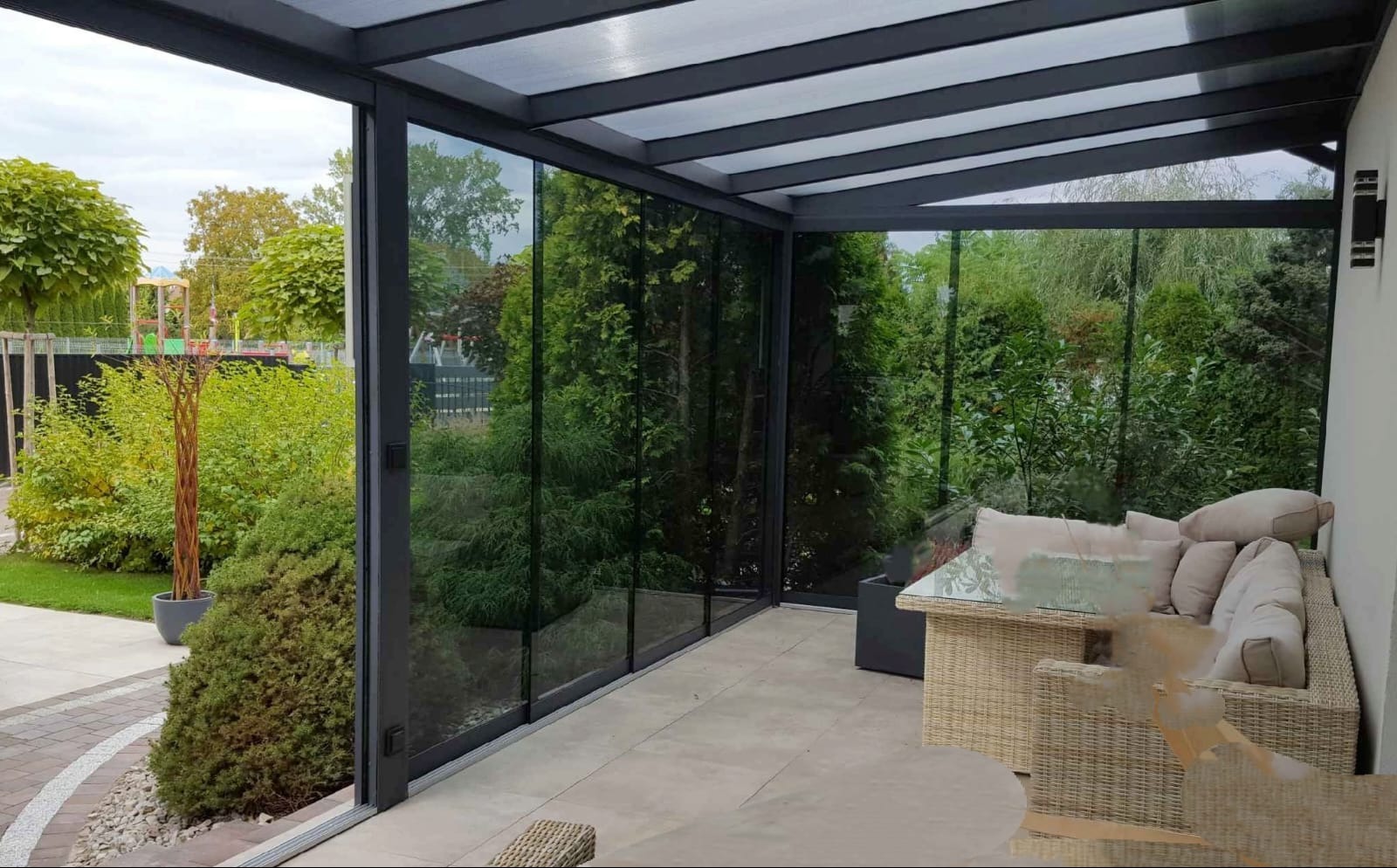 Zadaszony taras z przesuwnymi szklanymi panelami, widok na zadbany ogród z trawnikiem i drzewami, rattanowe meble wypoczynkowe z poduszkami.