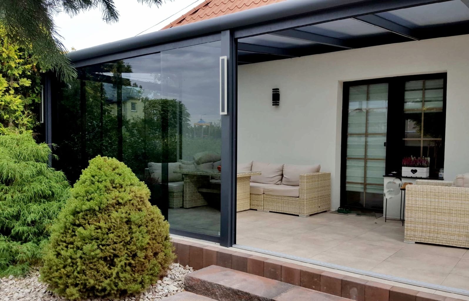 Zadaszony taras z przesuwnymi szklanymi ścianami, meble ogrodowe z rattanu, widok z zewnątrz na zadbany ogród.
