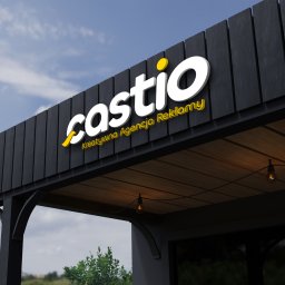 Castio.pl - Kreatywna Agencja Reklamowa i Drukarnia - Elewacja budynku z podświetlanym, trójwymiarowym logo 'castio' i napisem 'Kreatywna Agencja Reklamy' w kolorze żółtym, na tle błękitnego nieba.