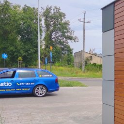 Castio.pl - Kreatywna Agencja Reklamowa i Drukarnia - Niebieski samochód kombi z reklamą firmy Castio, zaparkowany na chodniku obok nowoczesnego budynku z drewnianą elewacją.