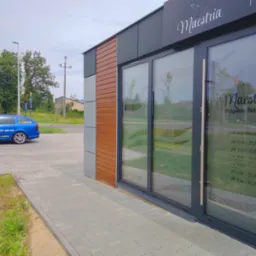 Fasada budynku z szyldem 'Maestria Przystań Relaksu' i godzinami otwarcia, obok zaparkowany niebieski samochód z reklamą firmy Castio.