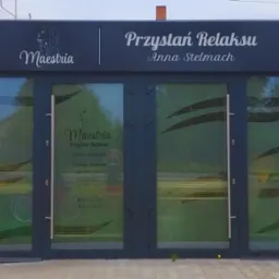 Fasada budynku z szyldem 'Przystań Relaksu Anna Stelmach' z logo Maeshia i godzinami otwarcia na naklejce na drzwiach, brązowe panele elewacyjne po bokach.