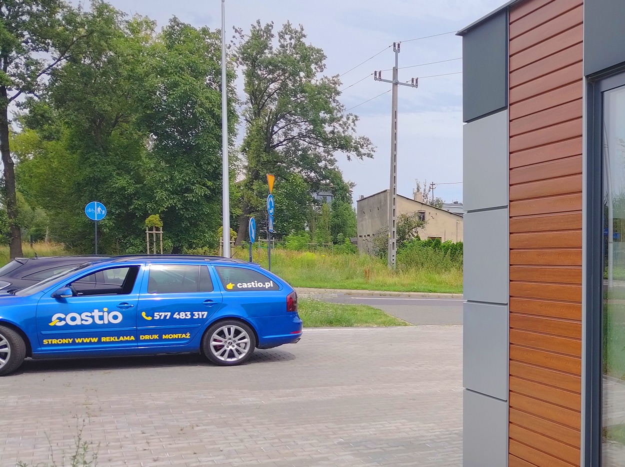 Niebieski samochód kombi z reklamą firmy Castio, zaparkowany na chodniku obok nowoczesnego budynku z drewnianą elewacją.