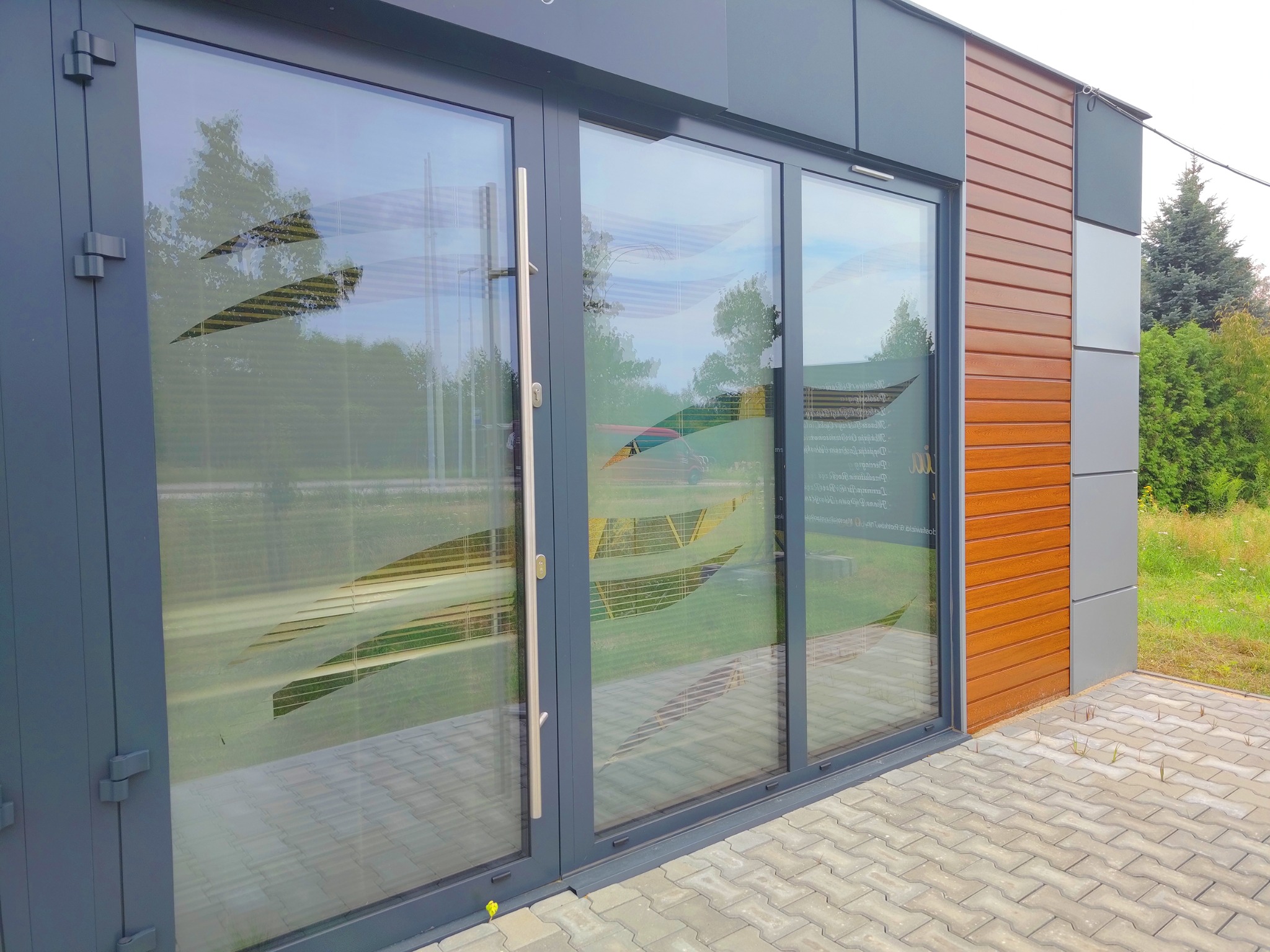 Szklane drzwi wejściowe z dekoracyjną folią imitującą żaluzje, widoczne z zewnątrz budynku o elewacji z paneli w odcieniach brązu i szarości.