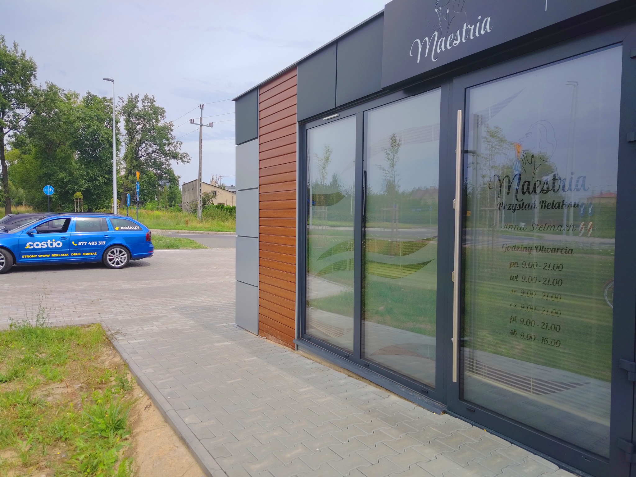 Fasada budynku z szyldem 'Maestria Przystań Relaksu' i godzinami otwarcia, obok zaparkowany niebieski samochód z reklamą firmy Castio.