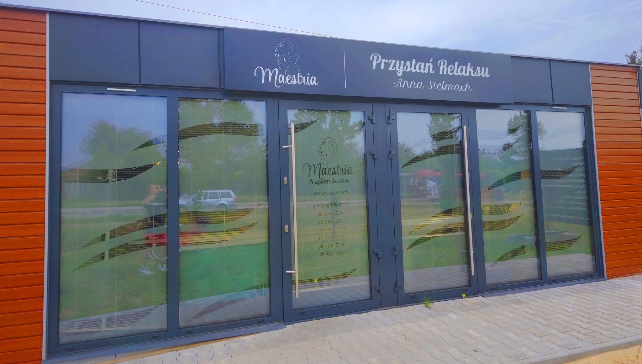 Fasada budynku z szarymi drzwiami wejściowymi i brązową okładziną drewnianą, nad wejściem szyld z nazwą 'Przystań Relaksu Anna Stelmach' i logo 'Maestria' z sylwetką kobiety.