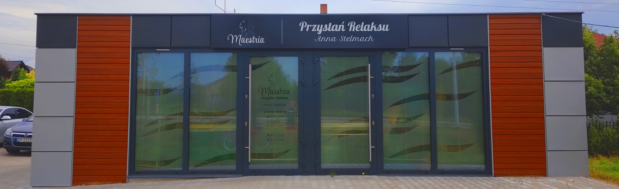 Fasada budynku z szyldem 'Przystań Relaksu Anna Stelmach' z logo Maeshia i godzinami otwarcia na naklejce na drzwiach, brązowe panele elewacyjne po bokach.