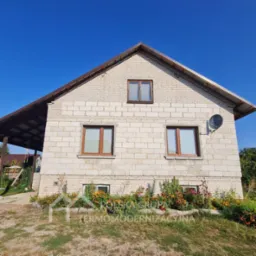 Dom z białej cegły z brązowymi oknami w Warszawie, widok z dołu, dach z blachy trapezowej, ogródek kwiatowy u podstawy elewacji, antena satelitarna.