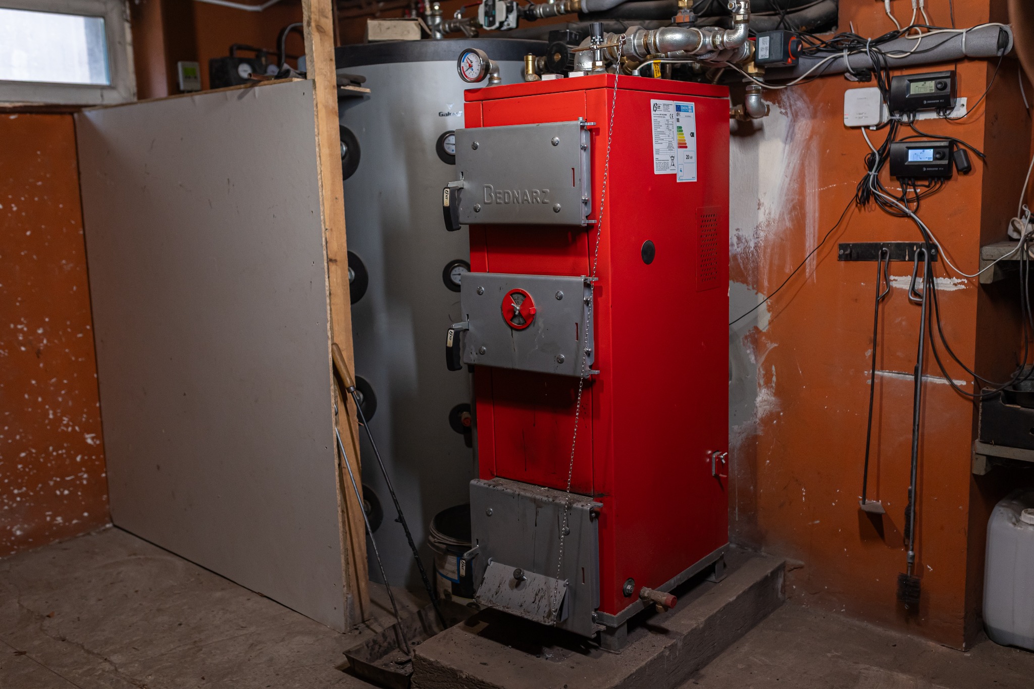 Czerwony piec Bednarz z szarymi drzwiczkami w kotłowni. Widoczne rury, manometry i regulatory. Pomarańczowa ściana z przewodami.