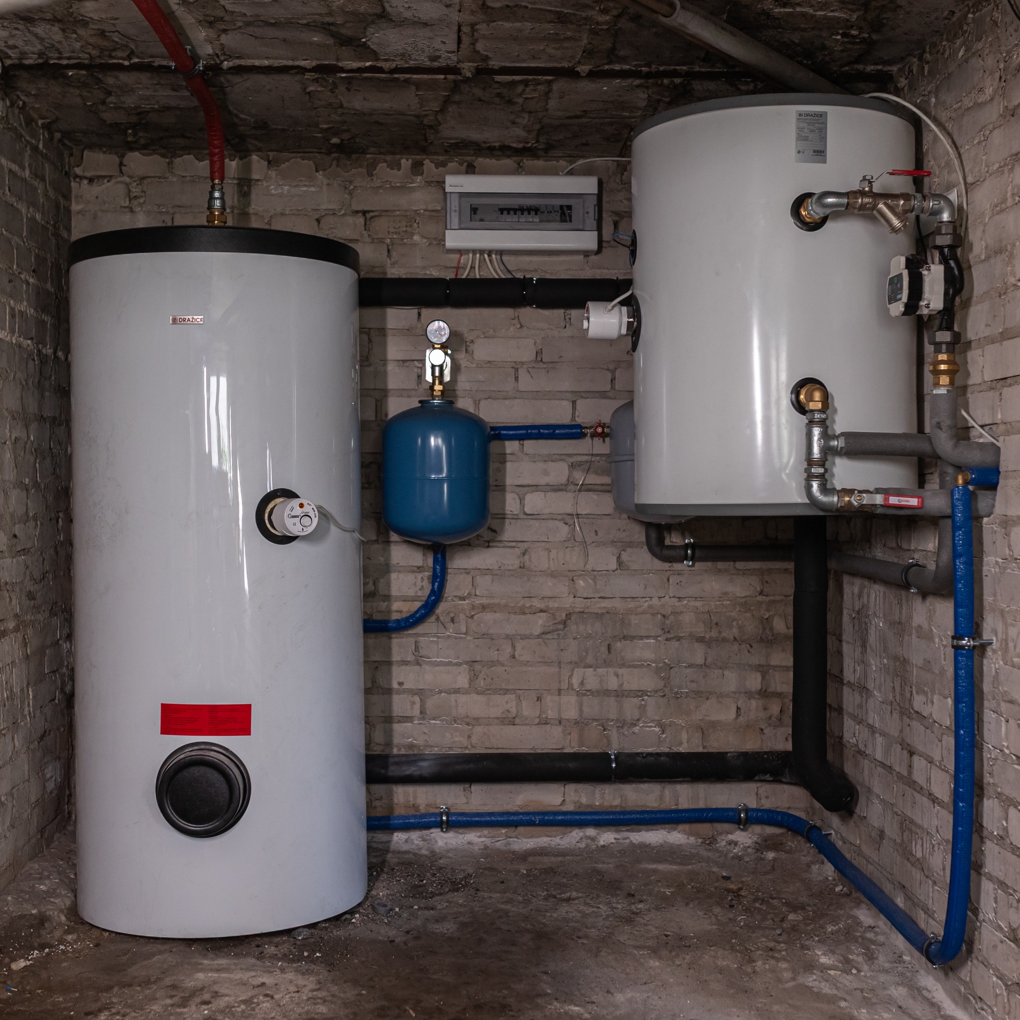 Dwa białe bojlery Drazice w piwnicy z ceglanymi ścianami, niebieskie rury i czarne izolowane rury, szara skrzynka elektryczna na ścianie.