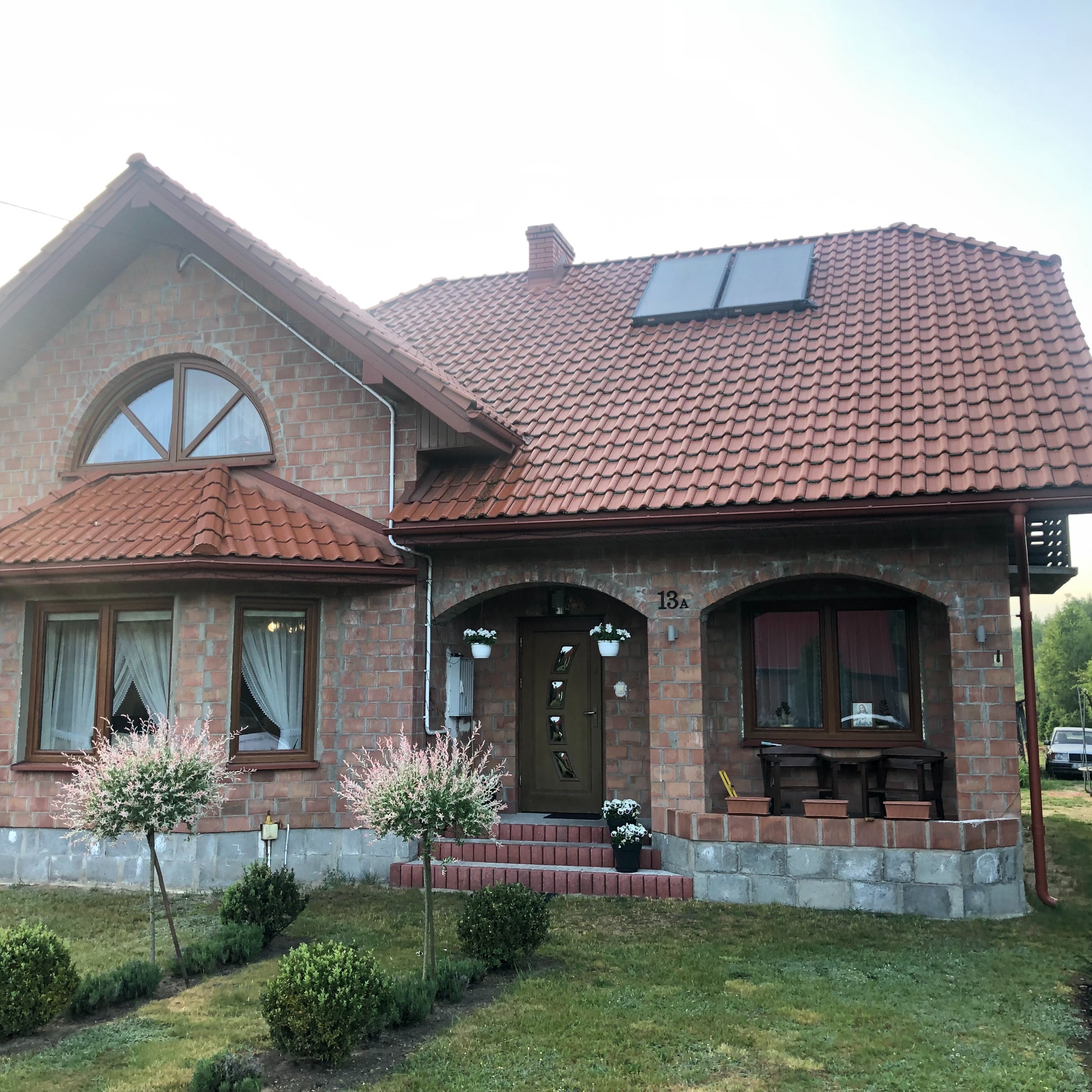 Dom z cegły klinkierowej z panelami słonecznymi na dachu, numerem 13A nad wejściem i dekoracyjnymi drzewkami w ogrodzie.