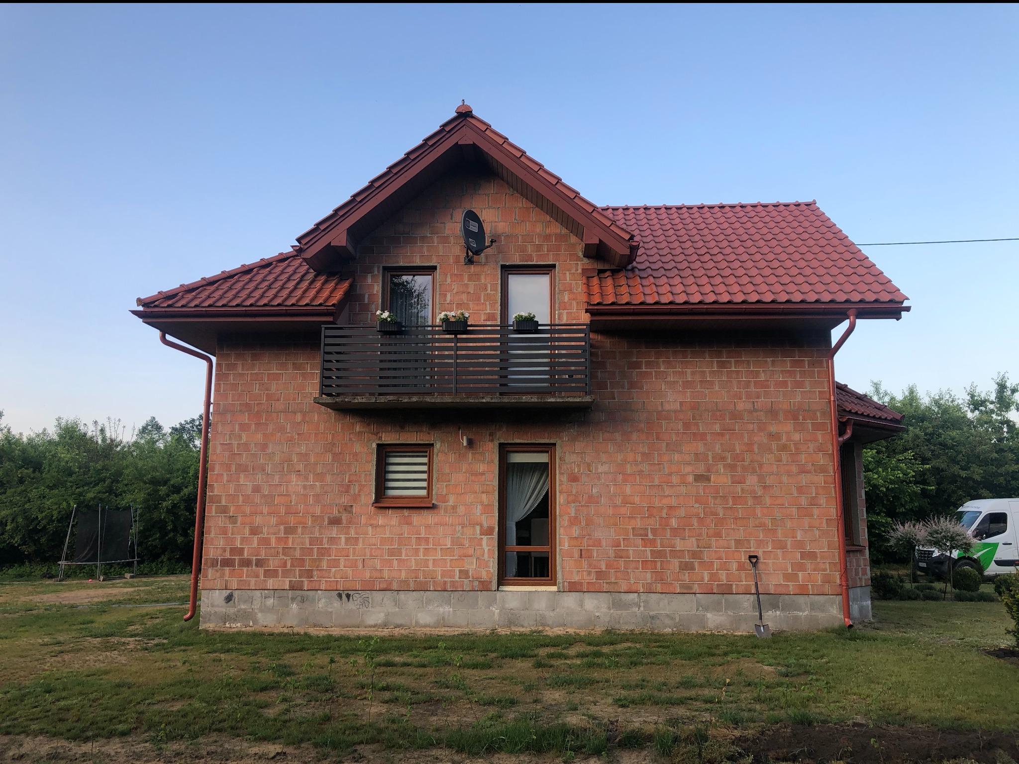 Dwukondygnacyjny dom z czerwonej cegły z balkonem, czerwonym dachem i instalacją satelitarną. Na trawniku widoczna łopata i samochód dostawczy.