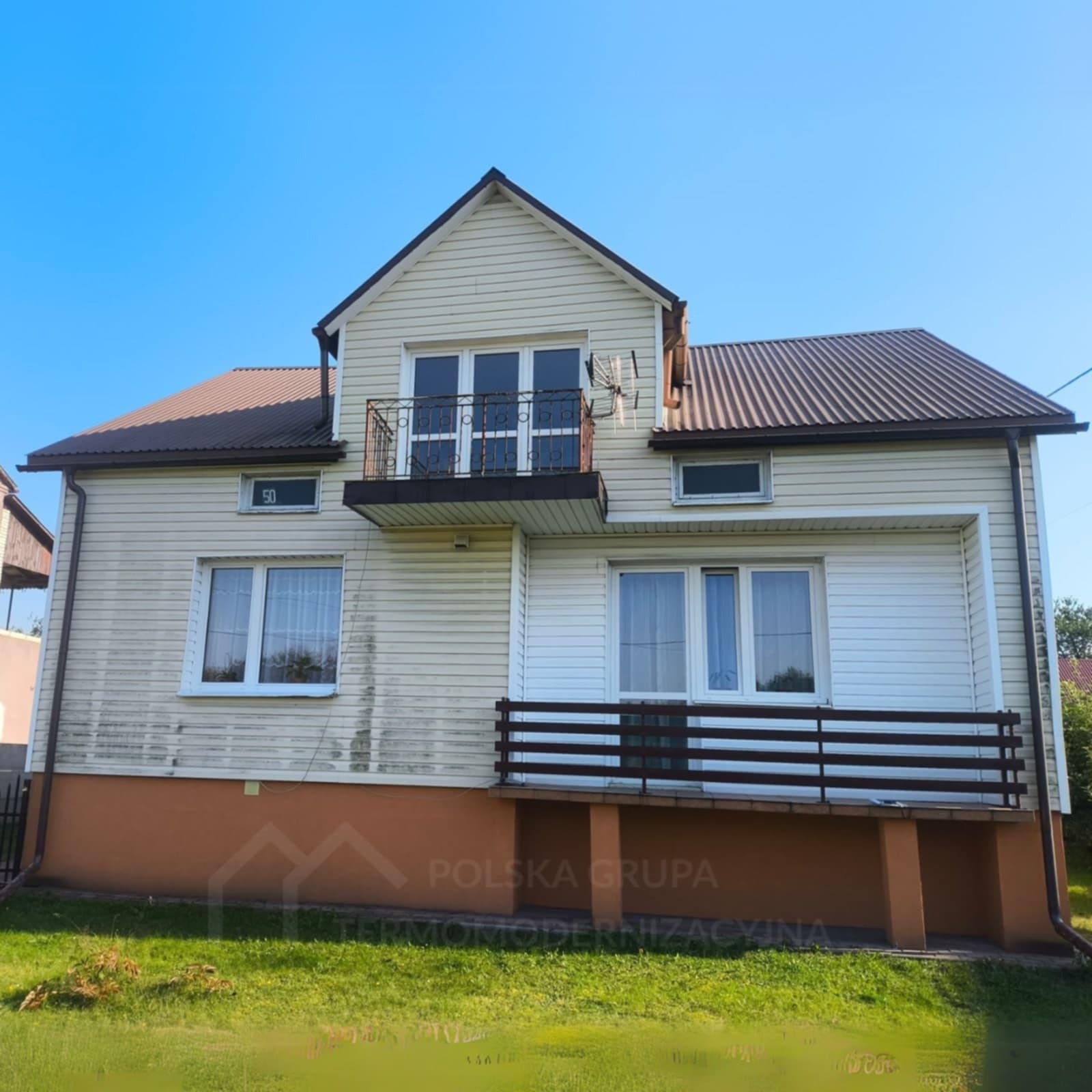 Dwupiętrowy dom z sidingiem i brązowym dachem, widziany z dołu, z wyraźnymi zabrudzeniami na elewacji i balkonem z kutą balustradą. Widoczne okna i brązowe rynny.