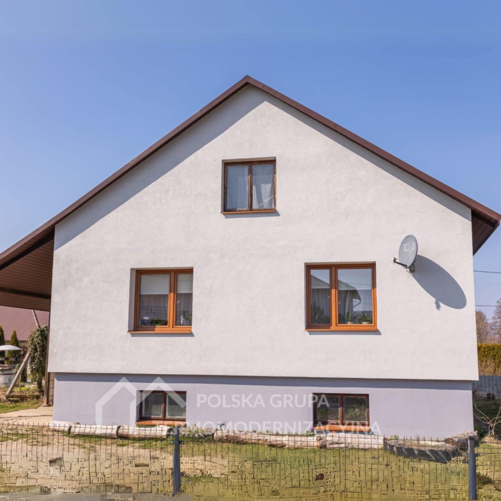 Dwukondygnacyjny dom z jasną elewacją i brązowymi oknami, widziany z przodu, z anteną satelitarną na ścianie. Ogród z metalowym ogrodzeniem na pierwszym planie.