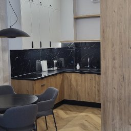 PKmeble - Nowoczesna kuchnia z drewnianymi szafkami dolnymi i białymi górnymi, czarnym blatem imitującym marmur, oświetleniem punktowym i podłogą ułożoną w jodełkę, widoczny stół z krzesłami.