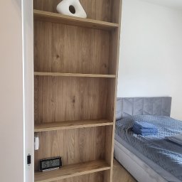 PKmeble - Wysoka, drewniana szafa z półkami w sypialni, na jednej z półek biały wazon dekoracyjny i elektroniczny zegarek, w tle widoczne łóżko z szarym zagłówkiem i niebieskimi ręcznikami.