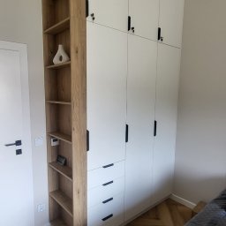 PKmeble - Wysoka, biała szafa z czterema górnymi drzwiczkami zamykanymi na klucz, trzema szufladami i otwartym regałem z drewnianymi półkami, uzupełniona wazonem i elektronicznym zegarkiem, na parkiecie...