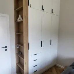 Wysoka, biała szafa z czterema górnymi drzwiczkami zamykanymi na klucz, trzema szufladami i otwartym regałem z drewnianymi półkami, uzupełniona wazonem i elektronicznym zegarkiem, na parkiecie...