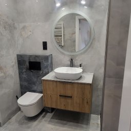 PKmeble - Nowoczesna łazienka z umywalką nablatową, szafką z drewnopodobnym frontem, okrągłym lustrem z podświetleniem LED i wiszącą toaletą, wykończona płytkami imitującymi marmur.