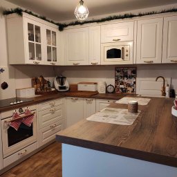 PKmeble - Biała kuchnia z drewnianymi blatami i złotymi uchwytami, ozdobiona świątecznymi girlandami i akcentami, widok na piekarnik z zawieszonymi ręcznikami w czerwoną kratę.