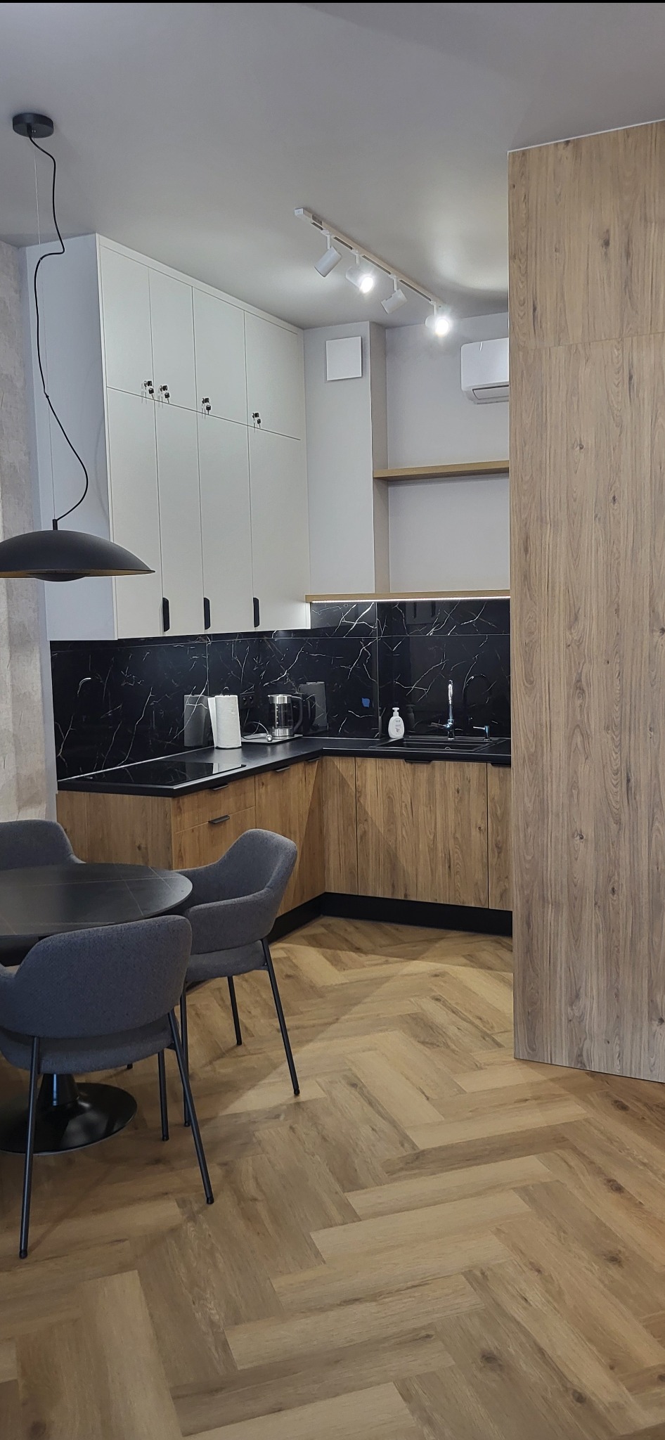 Nowoczesna kuchnia z drewnianymi szafkami dolnymi i białymi górnymi, czarnym blatem imitującym marmur, oświetleniem punktowym i podłogą ułożoną w jodełkę, widoczny stół z krzesłami.