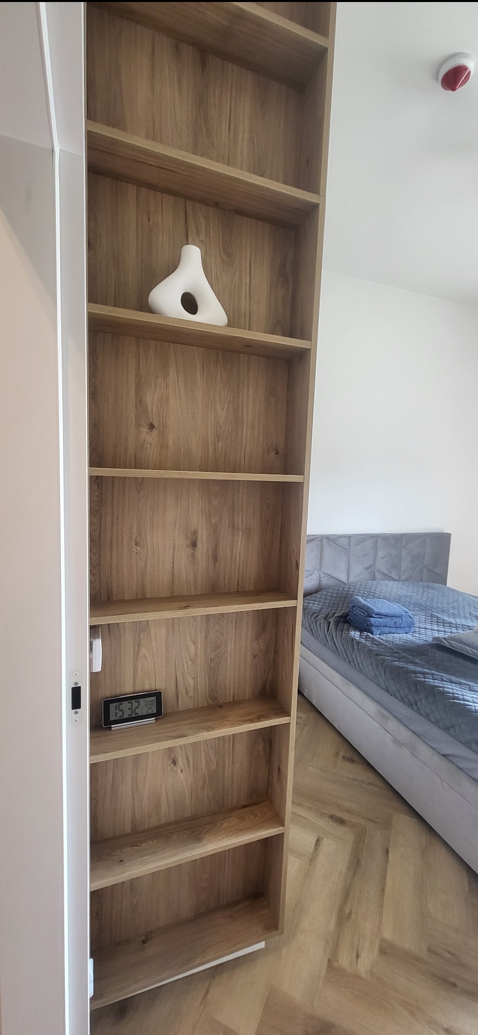 Wysoka, drewniana szafa z półkami w sypialni, na jednej z półek biały wazon dekoracyjny i elektroniczny zegarek, w tle widoczne łóżko z szarym zagłówkiem i niebieskimi ręcznikami.